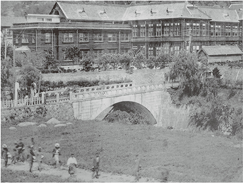 旧校舎の側面全景。長崎高等商業学校第八回卒業記念写真帖（1915（大正4）年）より。前方の石橋は1903（明治36）年に架設された拱橋（こまねきばし）（南西の方角から撮影）。学校の前にあって美観を損ねないよう、鉄骨トラス橋ではなく、アーチ橋が適当とされた。